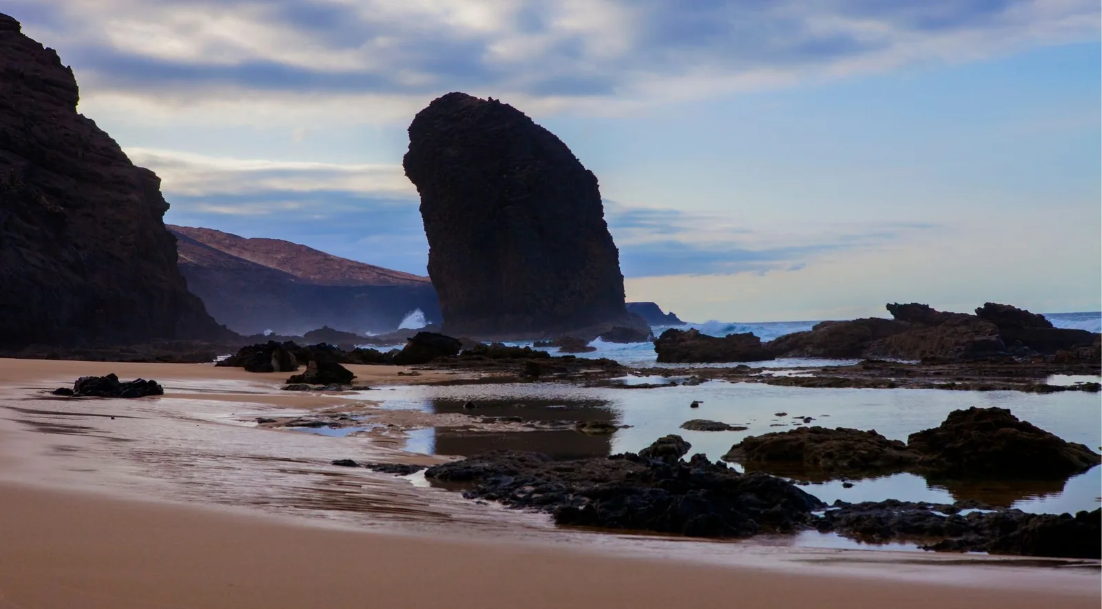 A large volcanic sea stack silhouetted at dusk above wet sand and tidal pools on a Lanzarote beach