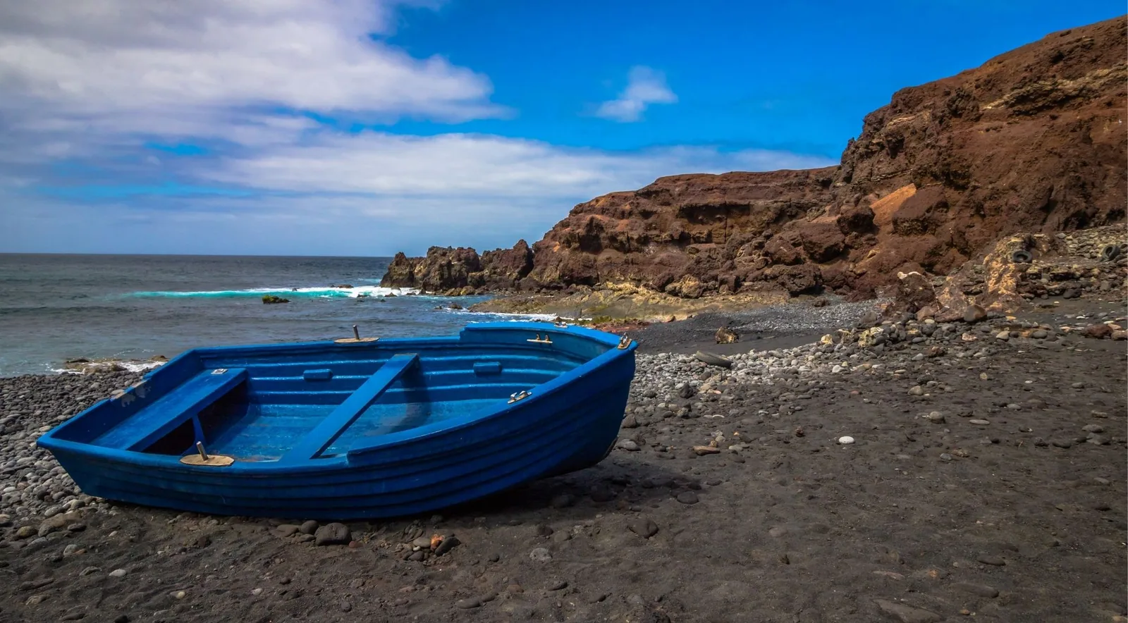A bright blue wooden fishing boat pulled up on a black pebble beach beneath red volcanic cliffs