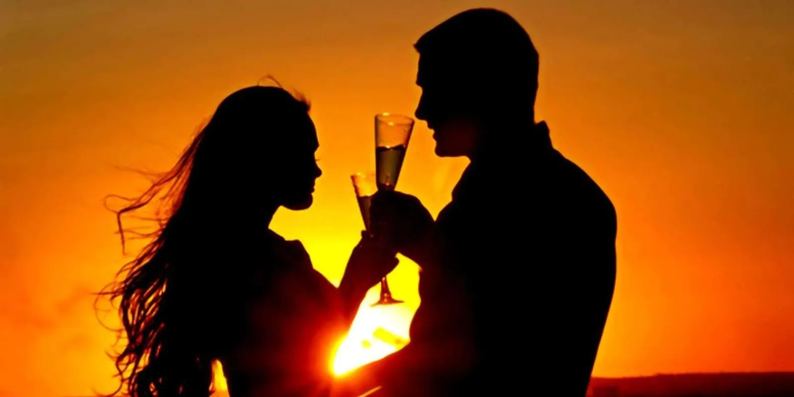 A couple silhouetted against a vivid orange sunset, toasting with sparkling wine on deck